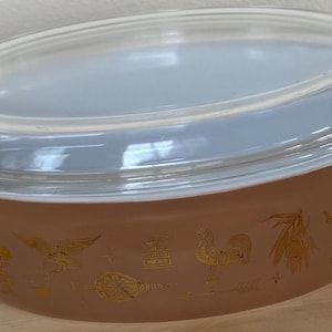 May include: An oval-shaped, tan Pyrex baking dish with a clear glass lid. The dish features gold-colored decorative images of a rooster, wheat, and other designs. The lid has a slight blue tint and fits snugly on the dish.