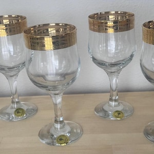 May include: Set of six clear wine glasses with gold rims. The glasses have a classic design with a rounded bowl and a slender stem. Each glass has a gold band around the rim and a small gold emblem on the base. The glasses are arranged on a light-colored wooden surface.