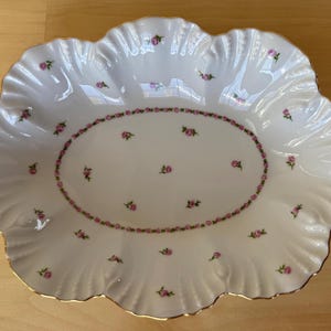 May include: A white oval serving dish with a scalloped edge and a gold rim. The dish is decorated with a delicate pattern of pink roses.