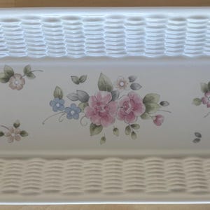 May include: Rectangular white ceramic basket-weave serving dish with floral design. The dish features a woven texture on the sides and a floral pattern with pink and blue flowers in the center. A decorative piece for serving.