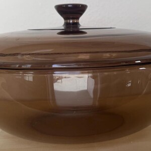 May include: A brown glass casserole dish with a matching lid. The dish has a rounded shape with two handles and a dark brown knob on the lid. The glass is transparent, allowing a view of the interior.