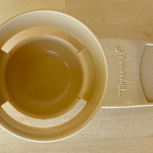 May include: A beige Tupperware egg separator. The separator has a round bowl with a slotted design and a handle with the Tupperware logo. The handle has a textured grip and a hole for hanging.