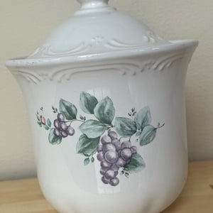 May include: White ceramic canister with a lid, featuring a painted design of purple grapes and green leaves. The canister has a decorative rim and a knob on the lid. A kitchen storage container.