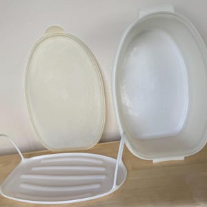 May include: A white oval Tupperware container with a matching lid and a white rack. The container is open, revealing its interior. The lid is off to the side. The rack has a handle on each side.