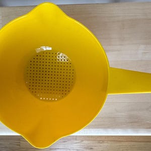 May include: A bright yellow plastic colander with a handle and a perforated straining surface. The colander has a wide bowl shape with a pouring spout and a small hole at the end of the handle. Kitchenware.