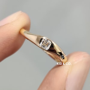 Op de afbeelding: Een gouden ring met een vierkant geslepen diamant in een bezel-zetting. De ring wordt tussen de vingers gehouden, wat de gepolijste afwerking benadrukt. De ring is van KIZI & CO.
