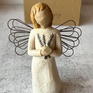May include: A carved wooden angel figurine with wire wings, holding a bundle of greenery. The angel has blonde hair and wears a cream-colored dress. The box behind the angel reads "Angel of Remembrance."