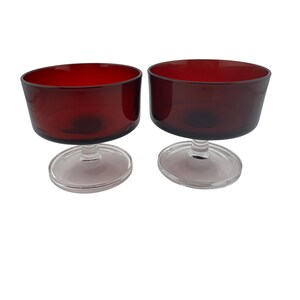 May include: A pair of matching dessert bowls featuring deep red glass basins set atop clear glass stems and circular bases. The bowls have a wide, open-top design with smooth, rounded edges, providing a consistent look for serving various food items.
