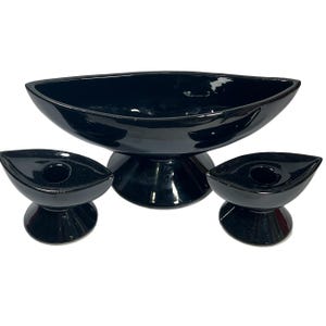 May include: A set of three black ceramic candle holders. The center holder is a large bowl with a wide base. The two smaller holders are shaped like boats with a small hole in the center for a candle.