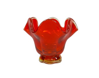 May include: A vibrant, orange-red glass bowl with a ruffled rim and a pedestal base. The bowl's design features a petal-like shape, and the base has a golden-yellow hue. The transparent glass allows light to pass through, highlighting the color.