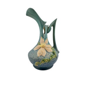 May include: A ceramic pitcher featuring a textured blue-green glaze with a prominent embossed cream-colored flower at the center. The vessel has a tall, flared spout, a curved handle, and green leaf accents extending from the base of the flower.
