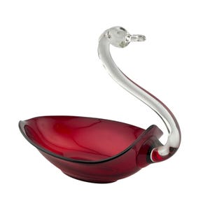 May include: A decorative glass swan-shaped bowl. The bowl is a deep red color with a clear glass swan neck and head. The swan's neck curves gracefully, forming a handle. The bowl is likely used for serving or display.