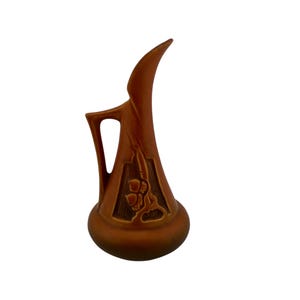 May include: A tall, rust-colored ceramic pitcher with a smooth, matte finish. The vessel features a wide, rounded base that tapers into a slender, elongated neck with a pointed spout. A single angular handle is attached to the side of the neck.