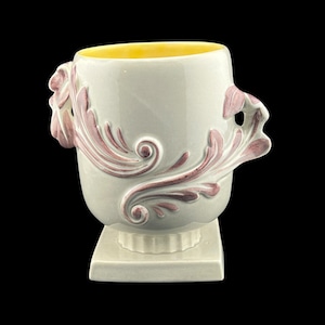 May include: A light gray ceramic vase with a yellow interior and pink floral relief design. The vase has two handles and sits on a square base. The vase is approximately 6 inches tall.