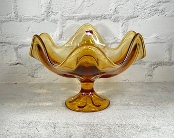 May include: Amber glass fruit bowl with a wavy rim and a pedestal base. The bowl is on a white textured surface.