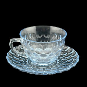 May include: A clear glass teacup and saucer set. The cup and saucer have a textured, bubble-like pattern. The cup has a handle and sits on a matching saucer with a scalloped edge. The set is against a black background.