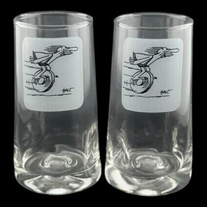 May include: Two clear glass tumblers with a cartoon image of a person riding a unicycle. The image is in black and white and is printed on a white rectangle on the front of each glass. The glasses have a flared shape.
