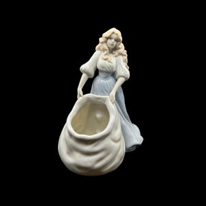 May include: A porcelain figurine of a figure with long blonde hair wearing a light blue dress with puffed sleeves. The figure is holding a large, white, sack-like vessel with a wide, open top. The piece features a smooth, glossy finish.