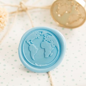 May include: A wrapped gift with a light blue wax seal depicting a world map. The gift is wrapped in white paper with a light green polka dot pattern and tied with twine. A wooden handled stamp is also visible.