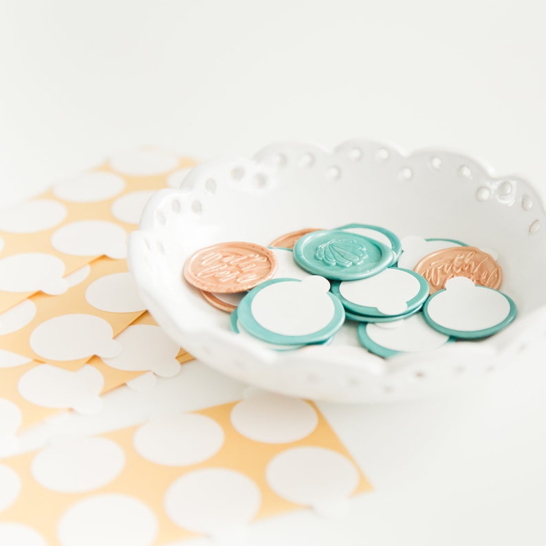May include: A white, scalloped-edge bowl filled with decorative wax seals in shades of teal, copper, and white. Some seals have seashell or script designs. A sheet of yellow paper with white polka dots is in the background.
