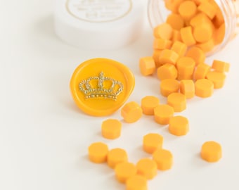 May include: A yellow wax seal with a silver crown design, surrounded by scattered orange wax beads. A clear plastic container and a white lid are also present, suggesting a crafting or stationery theme.