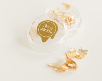 May include: A small clear plastic container with a gold lid and a gold sticker that says "Jessy Al Sea". The container is filled with gold leaf flakes. There are also some gold leaf flakes scattered on a white surface.