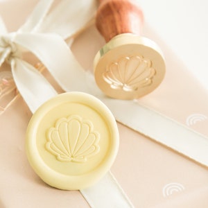 Könnte beinhalten: Ein gelbes Wachssiegel mit einem Muschelmotiv auf einem hellbraunen Hintergrund. Das Wachssiegel befindet sich neben einem goldenen Stempel mit einem Muschelmotiv.