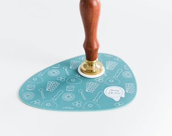 May include: A blue silicone mat with a white honeycomb pattern and the text "Jessy At Sea" in a circle. A wooden handled wax stamp with a gold base is on top of the mat.