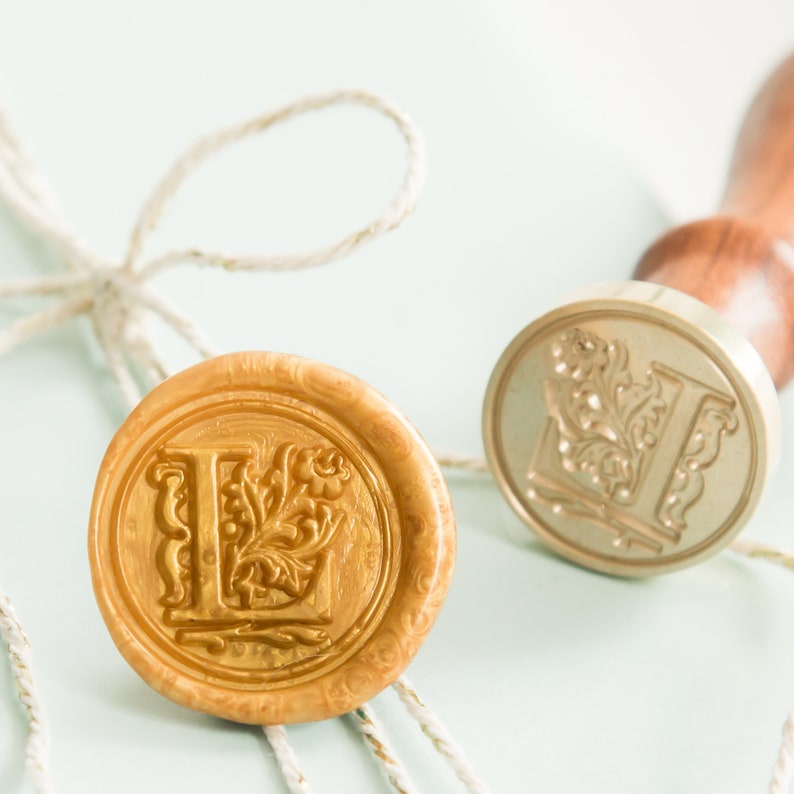 Op de afbeelding: Een gouden waszegel met de letter L en een bloemmotief. De zegel ligt op een wit oppervlak met een stuk touw.