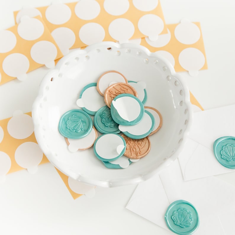 May include: A collection of teal and gold wax seals with a seashell design, some with adhesive backing, are displayed in a white, scalloped bowl. Several white envelopes with matching seals are also visible, along with sheets of white stickers on yellow backing.