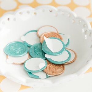 May include: A white, scalloped-edge bowl filled with teal and gold wax seals. Each seal has a white, peel-off adhesive backing. Some seals feature embossed floral designs. The background is a yellow surface with white polka dots.