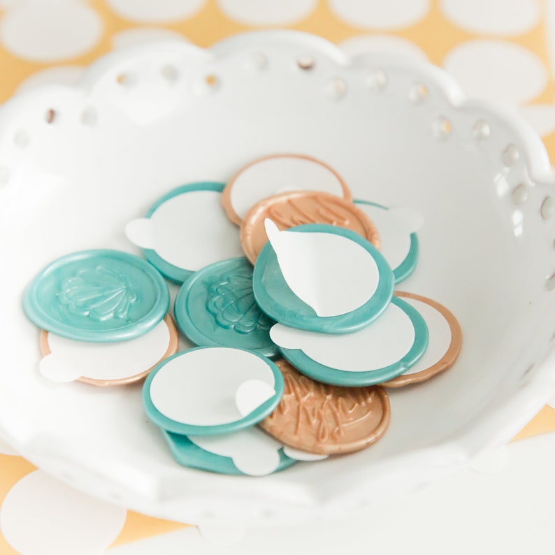 May include: A white, scalloped-edge bowl filled with teal and gold wax seals. Each seal has a white, peel-off adhesive backing. Some seals feature embossed floral designs. The background is a yellow surface with white polka dots.
