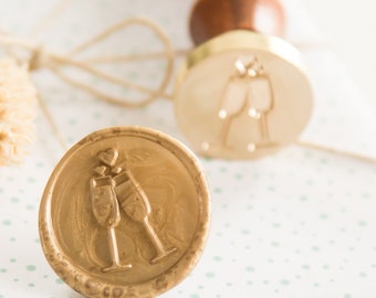 May include: A gold wax seal with a design of two champagne glasses and hearts. The seal is on a white background with green polka dots.