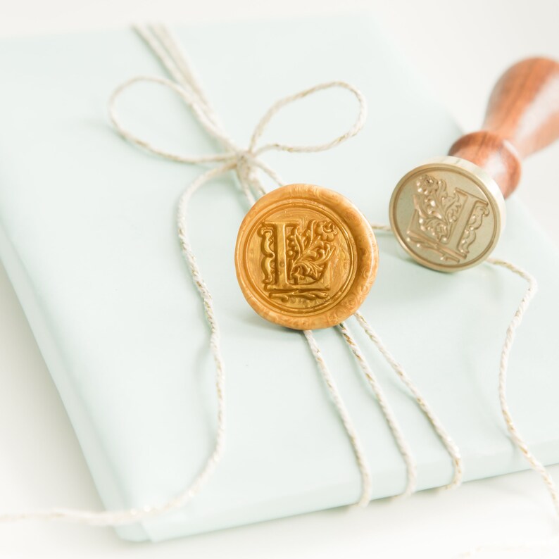 Op de afbeelding: Een lichtblauwe envelop met een gouden waszegel met de letter "L" en een decoratief ontwerp. De envelop is vastgebonden met een stuk touw.