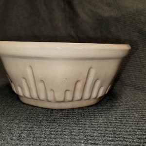 May include: A beige ceramic mixing bowl with a wide, flat rim and a series of vertical, recessed grooves decorating the lower exterior. The bowl features a smooth, matte finish and a rounded base, presenting a functional design for kitchen use.