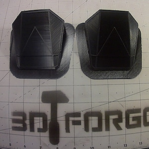 May include: Two black 3D printed geometric shapes with a raised triangular design. The shapes are on a gray grid surface with the text "3D FORGE" printed on it.