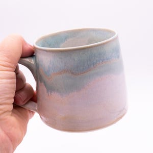 May include: A ceramic mug featuring a smooth, rounded silhouette with a handle on the side. The exterior displays a blended glaze finish, transitioning from a soft, pale blue at the top rim to a warm, muted pink and beige tone toward the base.