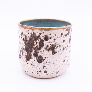May include: A ceramic mug with a speckled, off-white exterior and dark brown splatters. The rim has a brown band and a teal interior. The mug has a rounded shape and is suitable for hot or cold beverages.