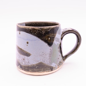 May include: A ceramic mug featuring a hand-applied glaze with abstract patches of dark brown, charcoal, and light lavender-grey. The mug has a cylindrical body, a curved handle on the right side, and a visible unglazed light tan base at the bottom.