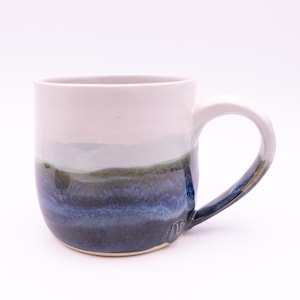 May include: A ceramic mug featuring a smooth white upper section that transitions into a blended gradient of earthy green and deep navy blue at the base. The handle is glazed in a matching dark blue tone, and a small stamped mark is visible near the base.