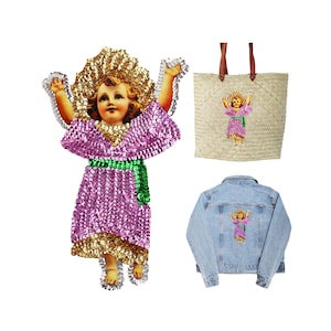 May include: A pink sequined angel with a gold halo and green sash. The angel is holding up its arms and has a smiling face. The angel is on a white background.