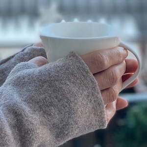 Könnte beinhalten: Eine weiße Keramiktasse mit Henkel wird von einer Person gehalten, die einen grauen Pullover trägt. Die Tasse scheint ein warmes Getränk zu enthalten, wobei Dampf von oben aufsteigt. Der Hintergrund ist unscharf, was auf eine Umgebung im Freien hindeutet.