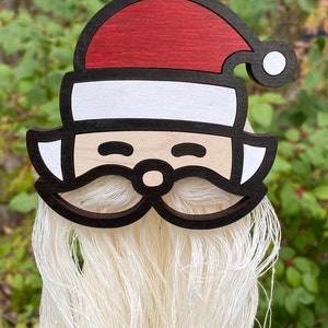 May include: A wooden Santa Claus ornament with a white yarn beard. The ornament is painted red and white and has a black outline. The ornament is hanging from a red and white polka dot ribbon.