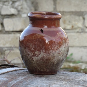 Puede incluir: Una olla de barro marrón con una superficie rugosa y texturizada. La olla tiene un pequeño agujero cerca de la parte superior y está sentada sobre una superficie de madera.