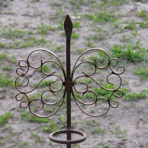 Pode incluir: Um suporte decorativo de metal enferrujado com um design de borboleta e um anel no topo. O suporte está sobre uma superfície de madeira branca.