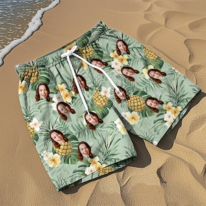 May include: Light green swim shorts featuring a repeating pattern of pineapples, tropical flowers, and a person's face. The shorts have an elastic waistband with white drawstring cords and a loose, relaxed fit for swimming or casual beach wear.