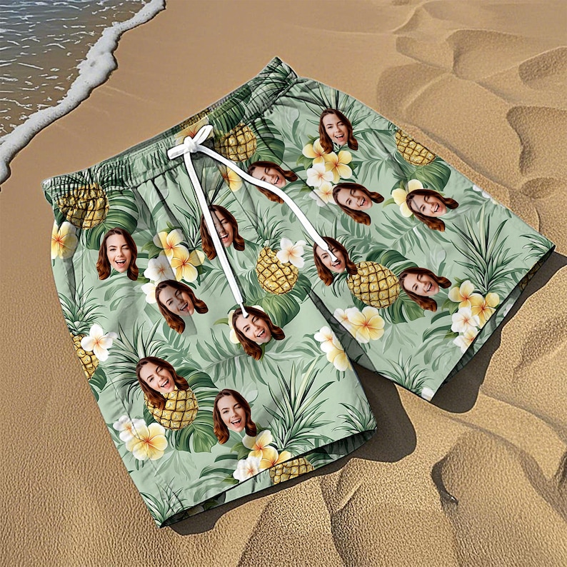 May include: Light green swim shorts featuring a repeating pattern of pineapples, tropical flowers, and a person's face. The shorts have an elastic waistband with white drawstring cords and a loose, relaxed fit for swimming or casual beach wear.
