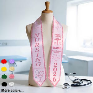 May include: A light pink nursing graduation sash with glitter trim, displayed on a mannequin. The sash features the word "NURSING", a stethoscope graphic, and the text "Class of 2026". A stethoscope rests nearby.