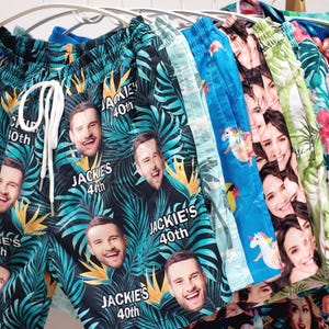 May include: A collection of swim trunks hanging on a rack, featuring various custom prints. The front pair is black with a tropical leaf pattern, repeating faces, and the text "JACKIE'S 40th". Other pairs display blue unicorn patterns, floral designs, and additional custom face prints.