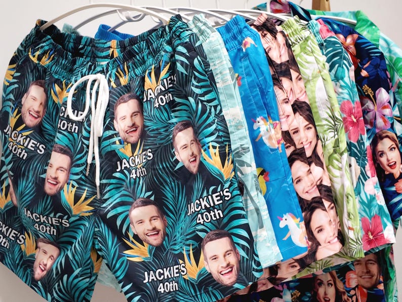 May include: A collection of swim trunks hanging on a rack, featuring various custom prints. The front pair is black with a tropical leaf pattern, repeating faces, and the text "JACKIE'S 40th". Other pairs display blue unicorn patterns, floral designs, and additional custom face prints.